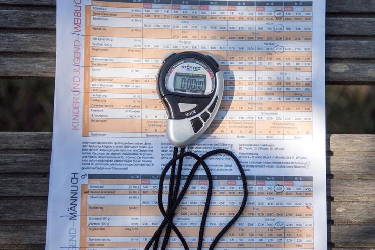 Stoppuhr und Infoblatt des Deutschen Sportabzeichens auf einer Holzbank im Freien.
