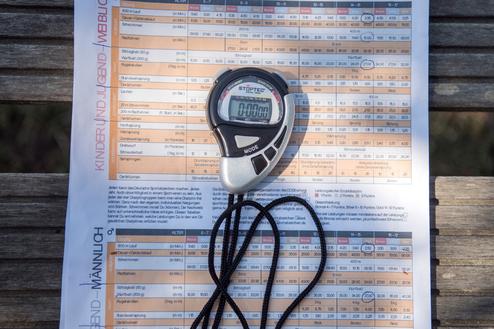 Stoppuhr und Infoblatt des Deutschen Sportabzeichens auf einer Holzbank im Freien.