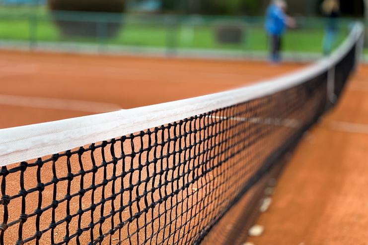 Nahaufnahme eines Tennisnetzes auf einem roten Sandplatz, Hintergrund unscharf mit grünen Bäumen.
