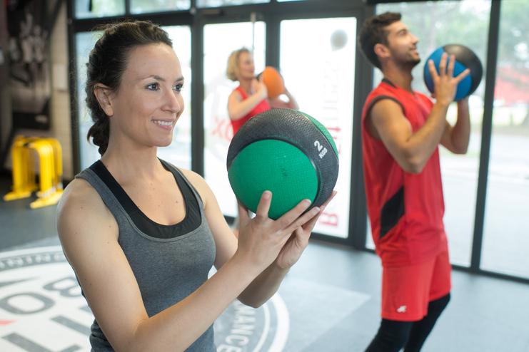 Drei Personen trainieren im Fitnessstudio mit Trainingsbällen; eine Frau hält einen grünen Medizinball.