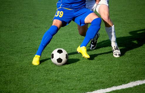 Zwei Fußballspieler kämpfen um den Ball auf einem grünen Kunstrasenplatz.
