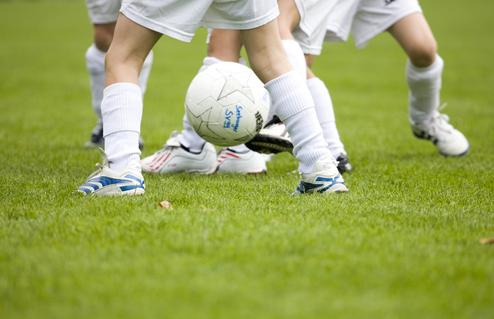 Spielende Kinder mit Fußball auf einem grünen Rasenplatz, nur Beine und Ball sichtbar.