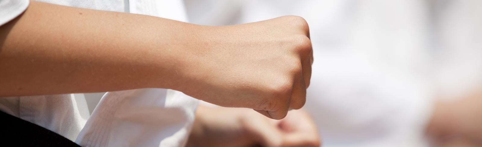 Nahaufnahme einer geballten Faust in einem weißen Karateanzug, im Hintergrund verschwommene Hände weiterer Teilnehmer.