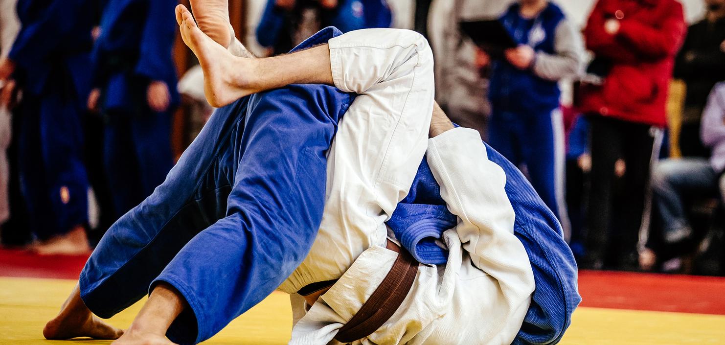 Zwei Judoka im Wettkampf auf einer gelben Matte, einer mit weißem, der andere mit blauem Gi, in einer Bodenkampfposition.