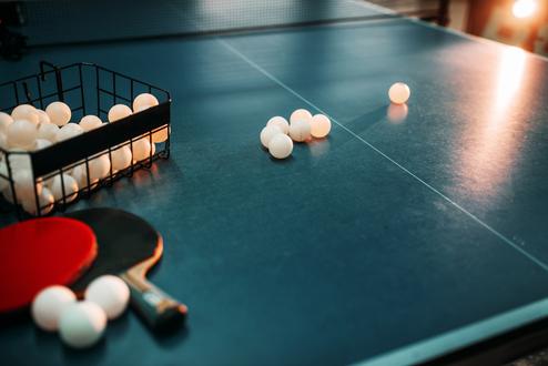 Nahaufnahme eines Tischtennisschlägers neben einem Korb mit Tischtennisbällen auf einer blauen Tischtennisplatte.