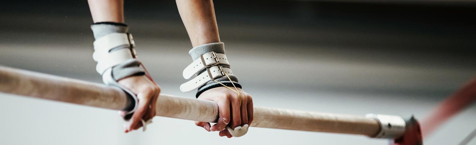 Nahaufnahme zweier Hände, die eine schmale Turnstange mit Griffpolstern halten, im Hintergrund eine Turnhalle.