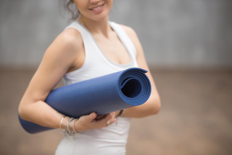 Frau hält eine gerollte blaue Yogamatte und lächelt, sie trägt ein weißes Top in einem Trainingbereich.